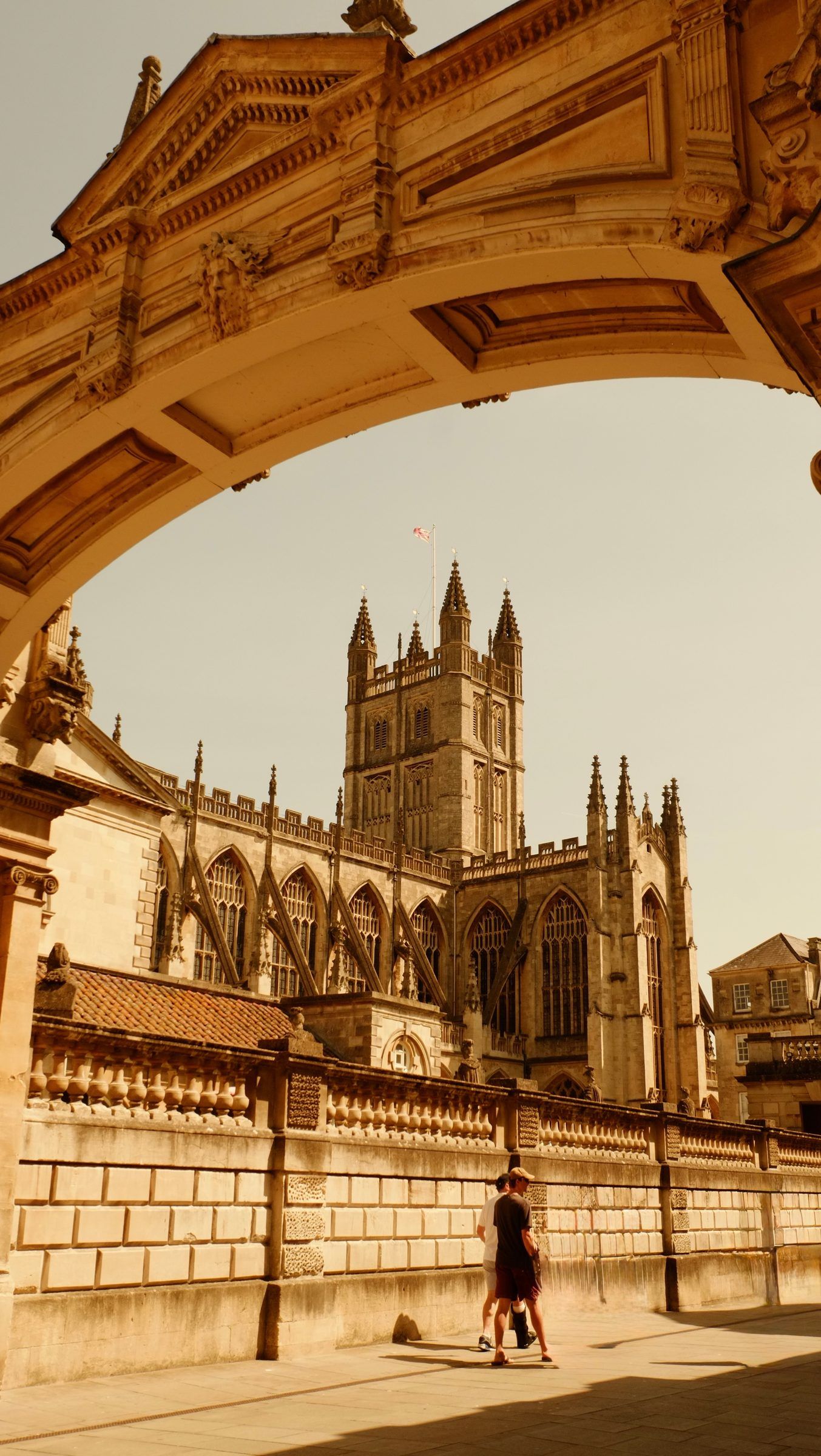 Bath Abbey's tower seen beneath a carved stone arch, two people walking past a balustraded wall in the sun