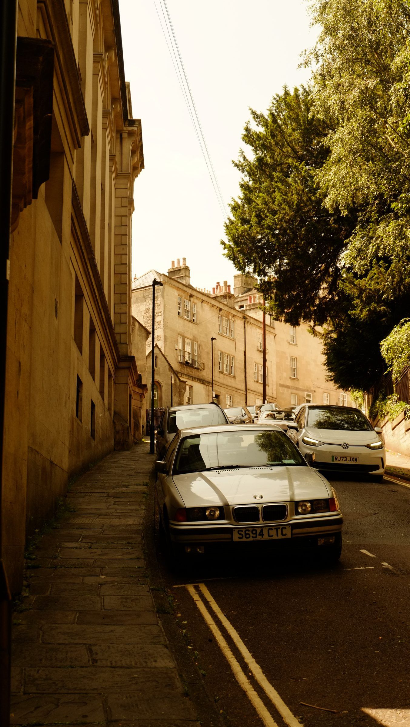 An old silver saloon parked on a steep, narrow lane between a stone wall and leafy trees