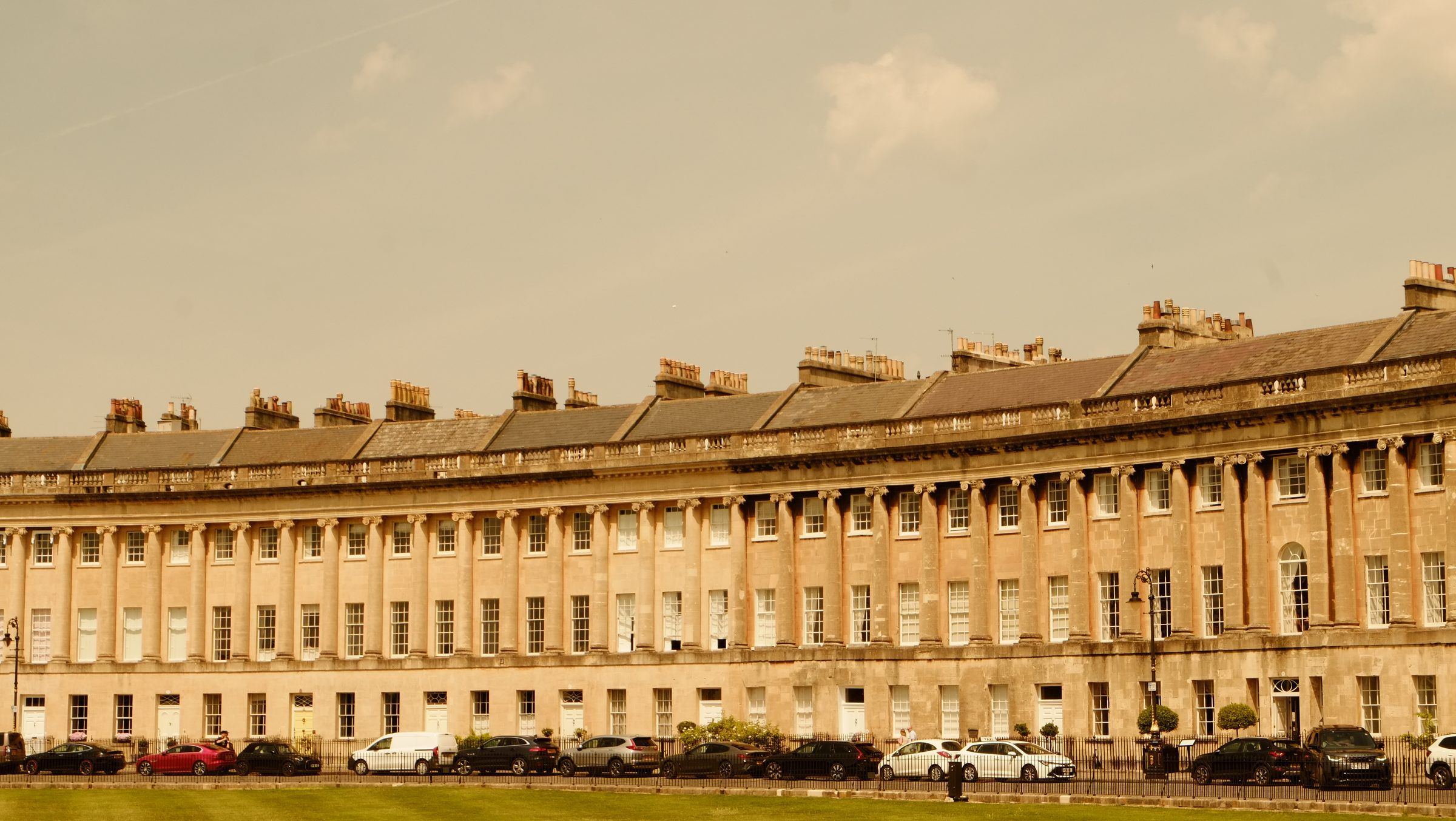 The columned front of the Royal Crescent with cars parked along the lawn's edge under a hazy sky