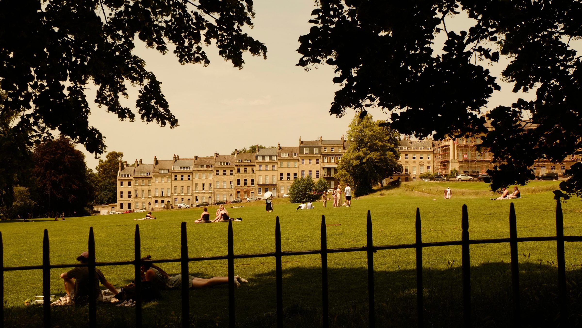 People lying on the grass in a park, framed by dark tree branches and a line of iron railings, townhouses beyond