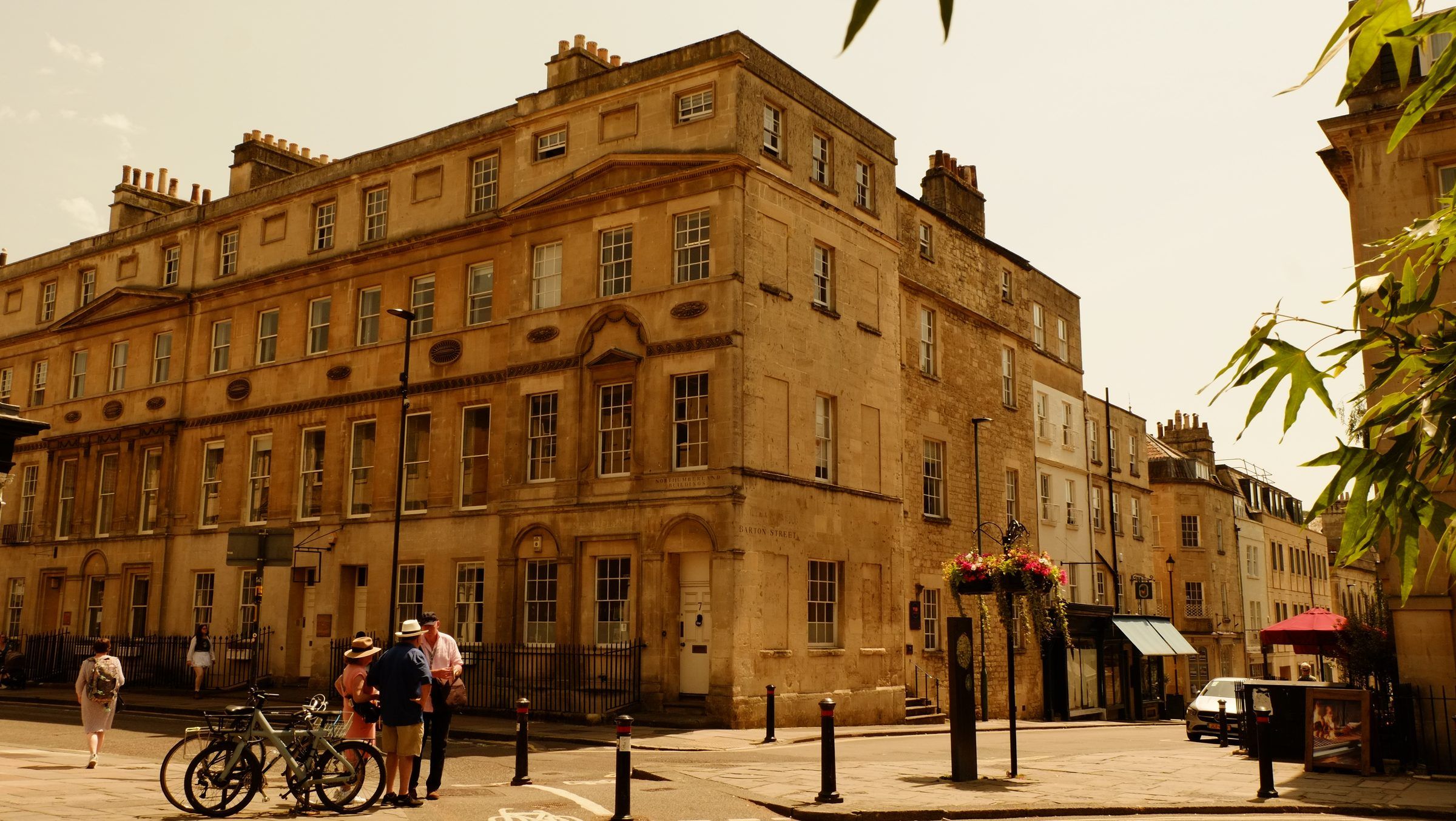 People chatting beside parked bicycles in front of a large Georgian corner building, hanging flower baskets nearby
