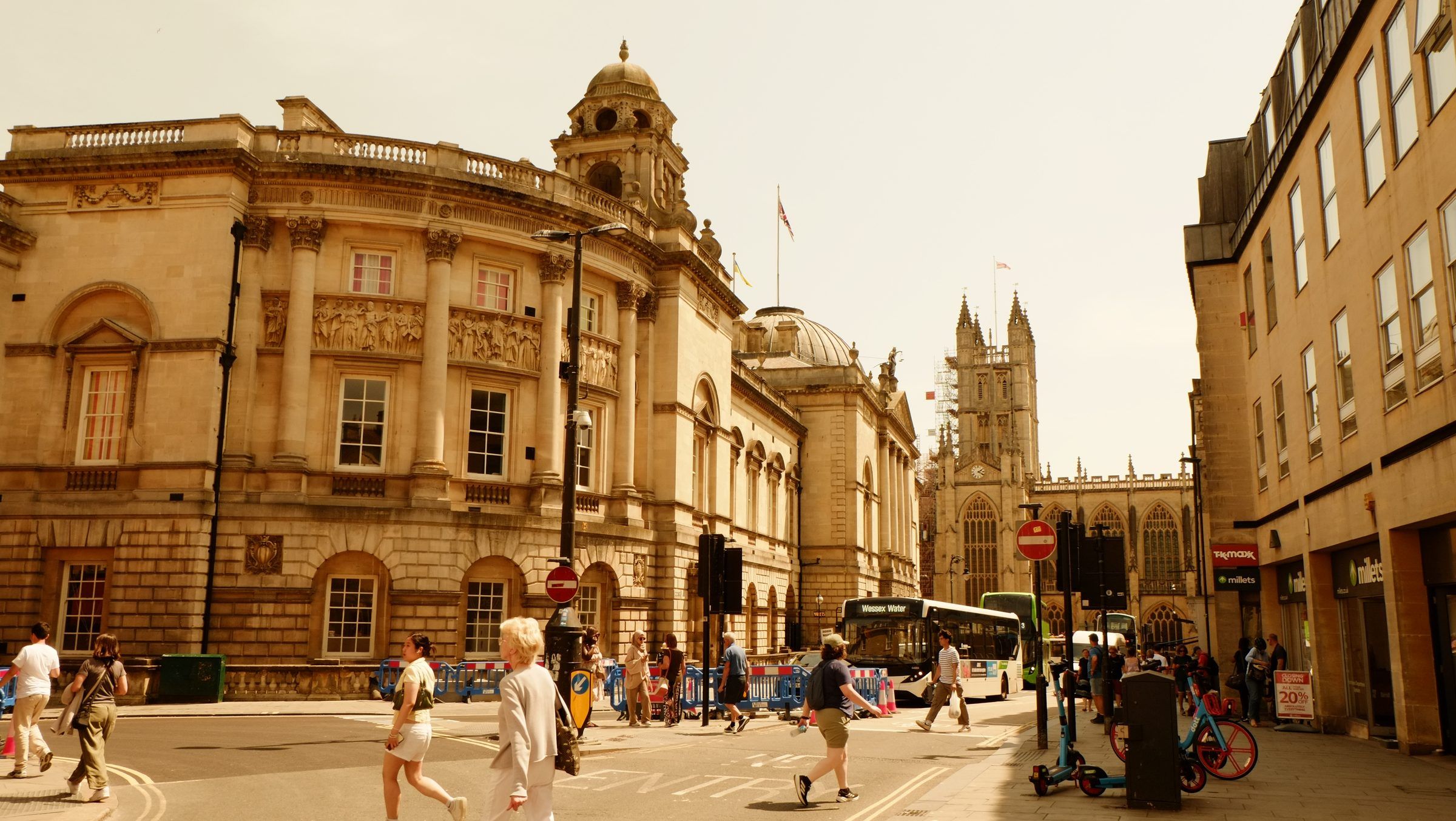 Pedestrians crossing in front of the domed Guildhall, a bus waiting and Bath Abbey at the end of the street