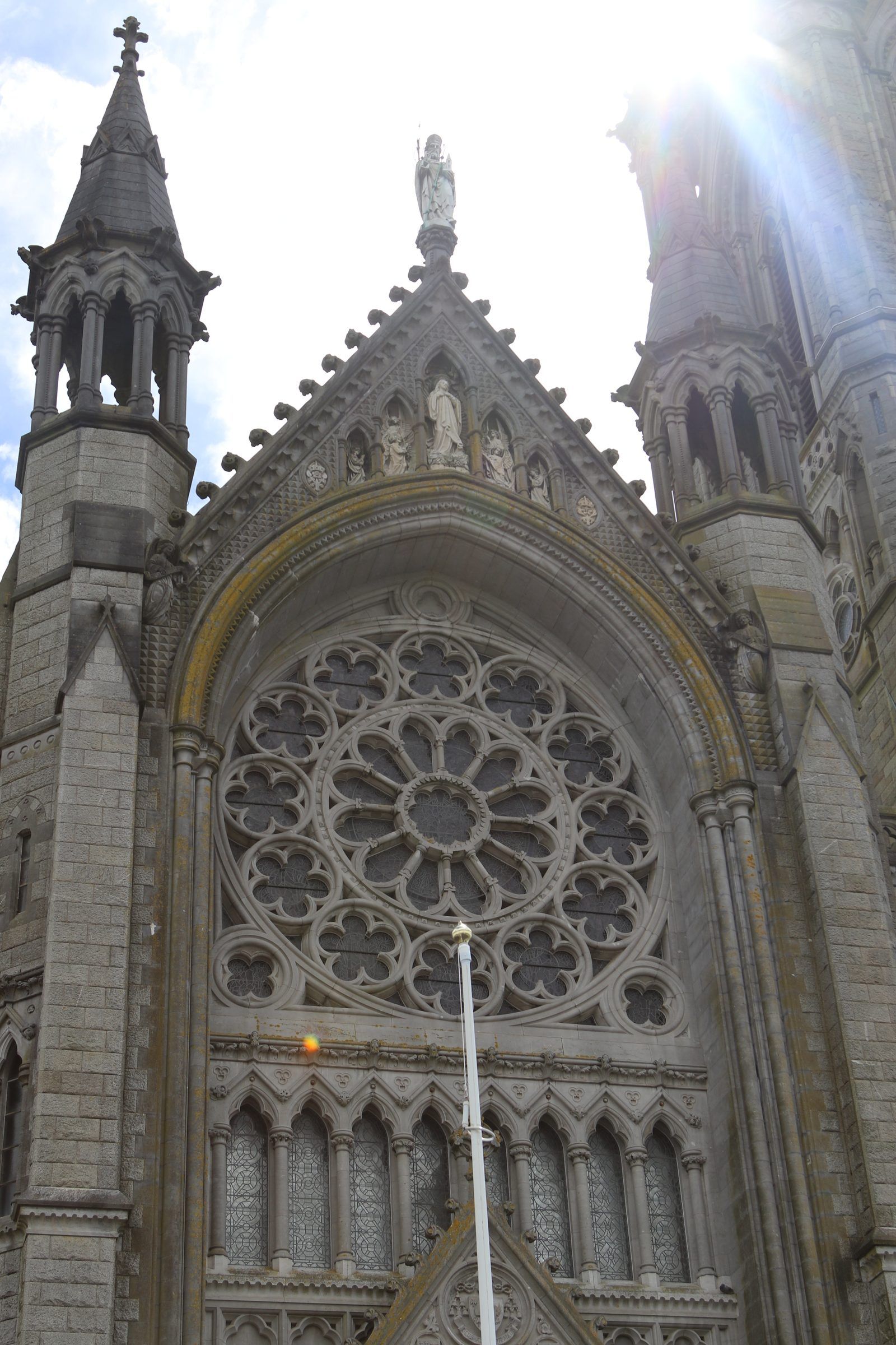Looking up at a Gothic cathedral front with a large rose window and spired turrets, sunlight flaring over the roof