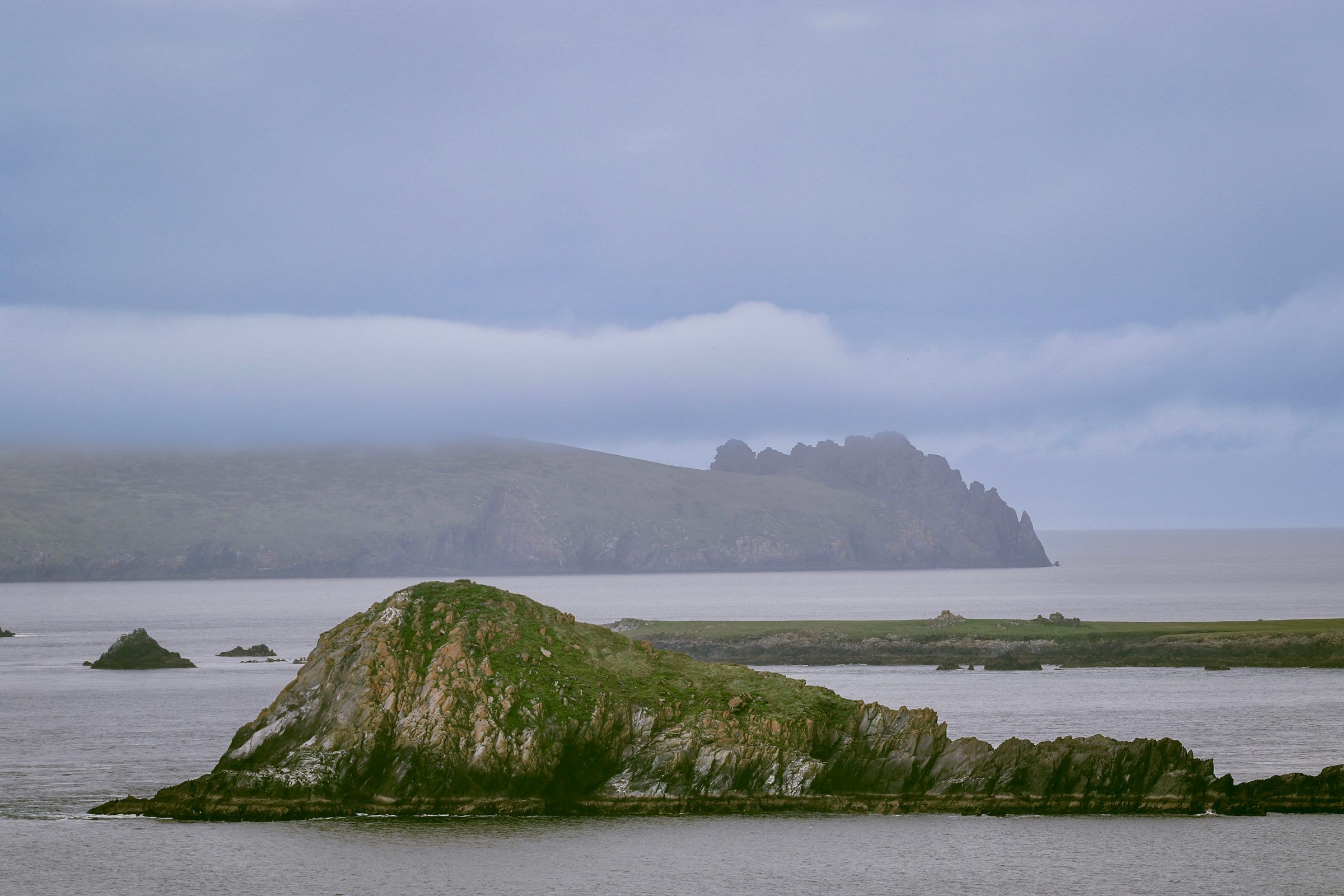 A grassy rock islet in a grey sea with a jagged headland silhouetted in the haze behind