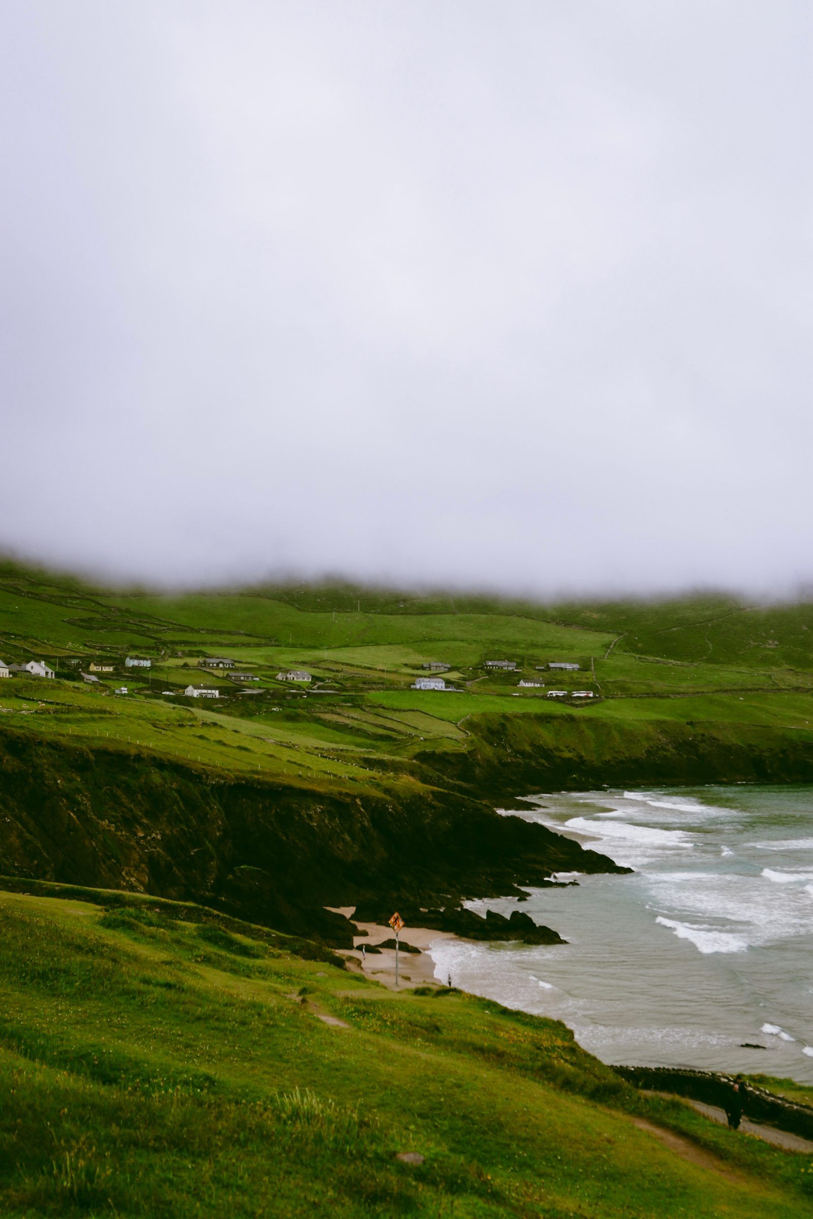 A sandy cove with breaking waves beneath green cliffs, white farmhouses scattered across the hillside in mist