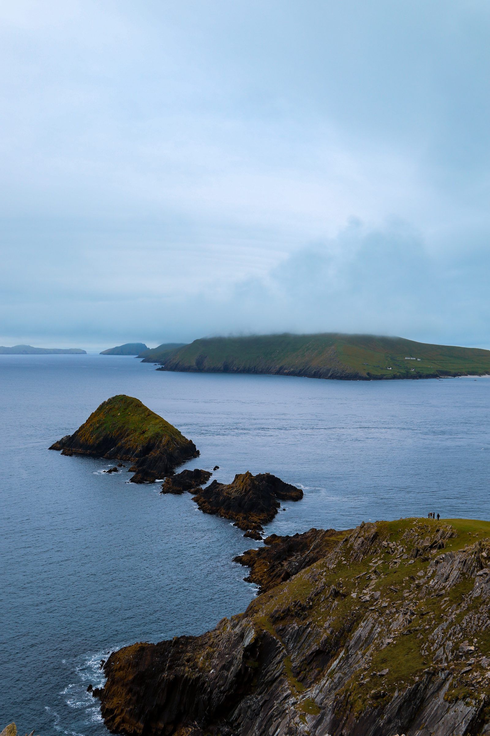 Rocky islets trailing off a headland into a calm blue sea, a long island under cloud on the horizon