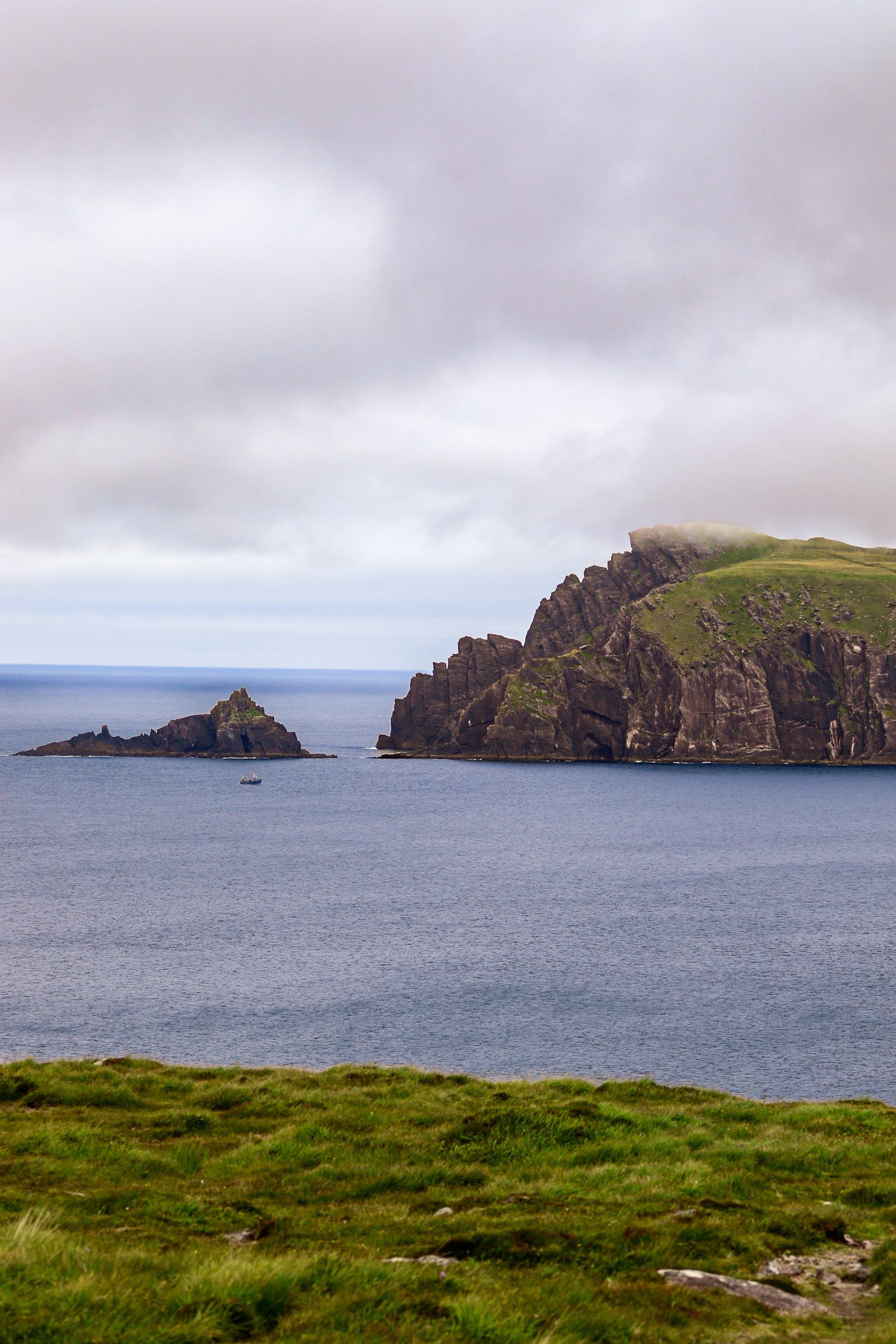 Dark sea cliffs and a rocky stack rising from a calm sea under pink-tinged cloud, a small boat passing between them