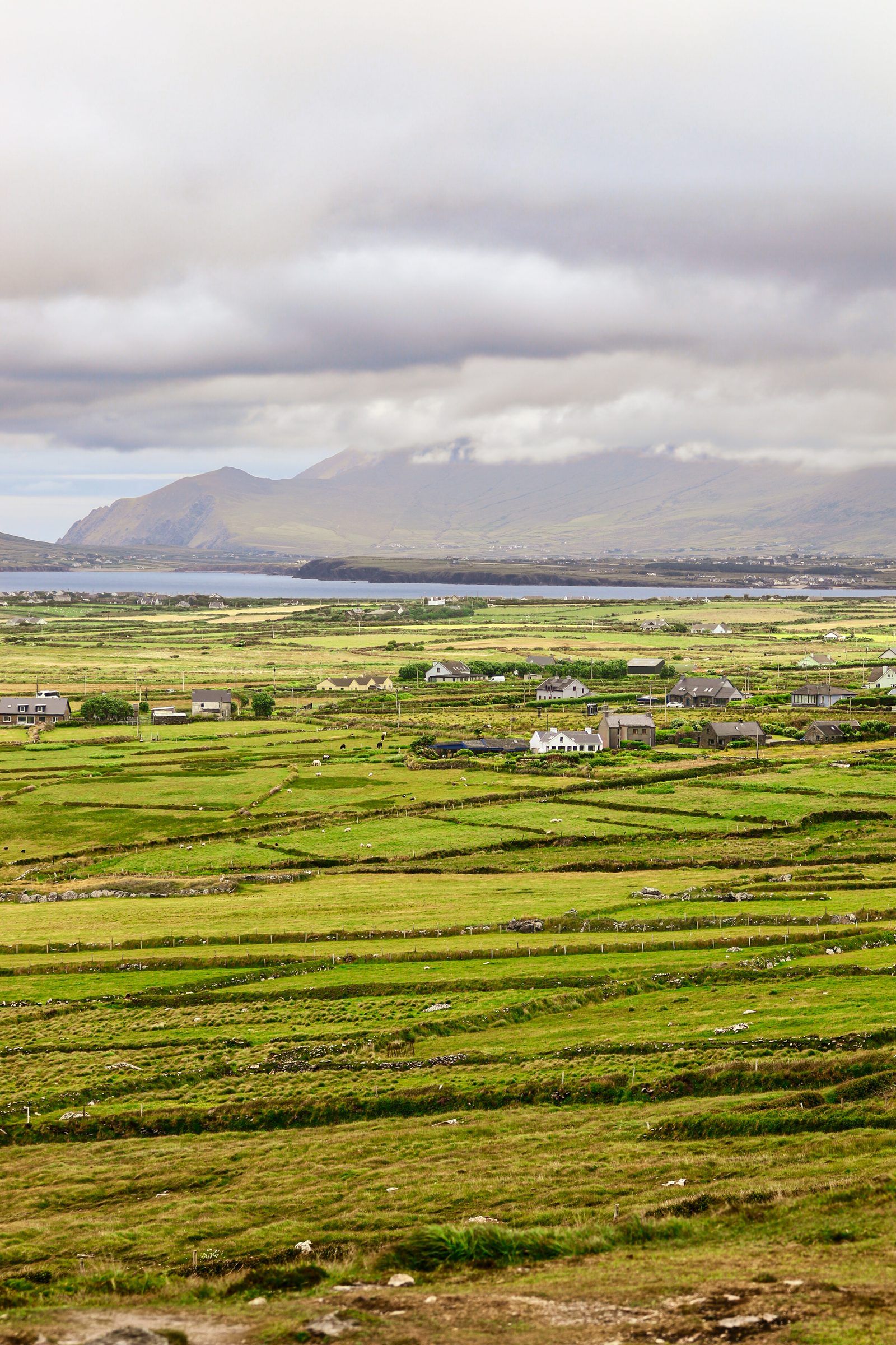 A patchwork of green fields and cottages stretching to a bay, mountains wrapped in cloud beyond