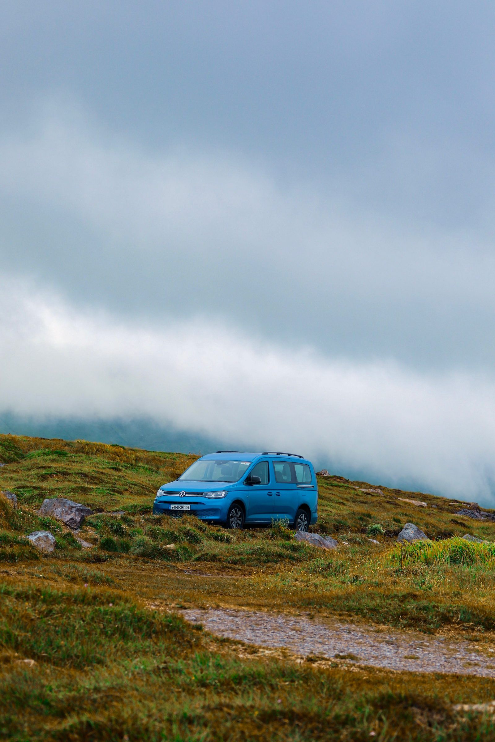 A blue campervan parked on a grassy hillside beneath a rolling bank of cloud