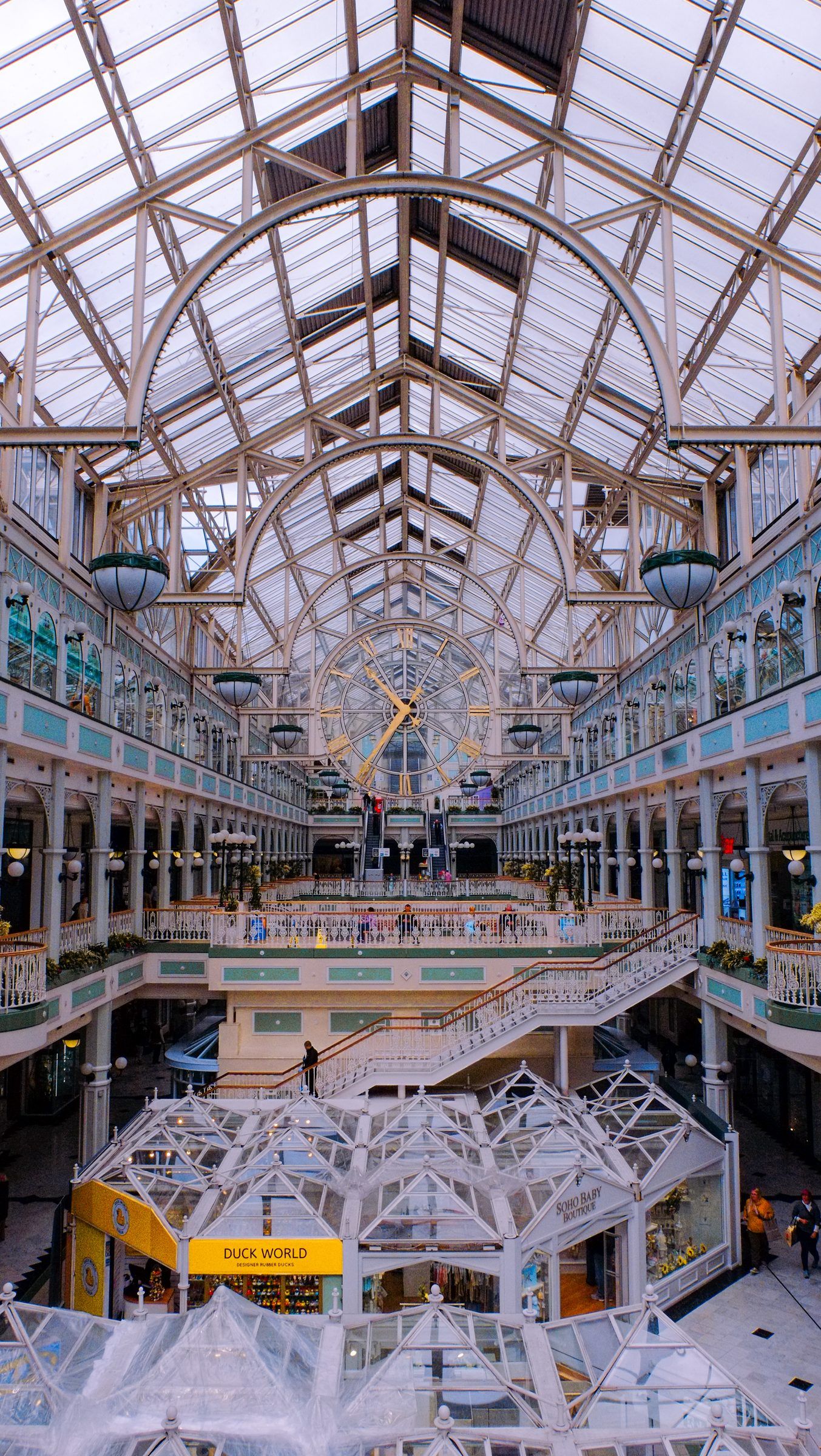 The white iron and glass roof of a shopping arcade with a large clock, balconies and a staircase below