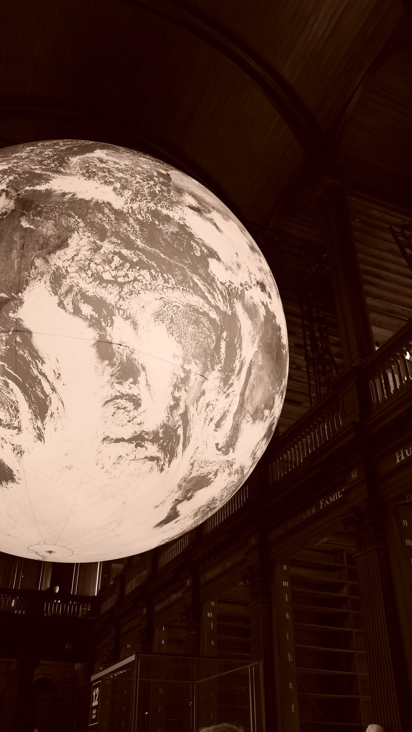 A sepia close view of a giant globe of the Earth suspended beneath the vaulted ceiling of an old library