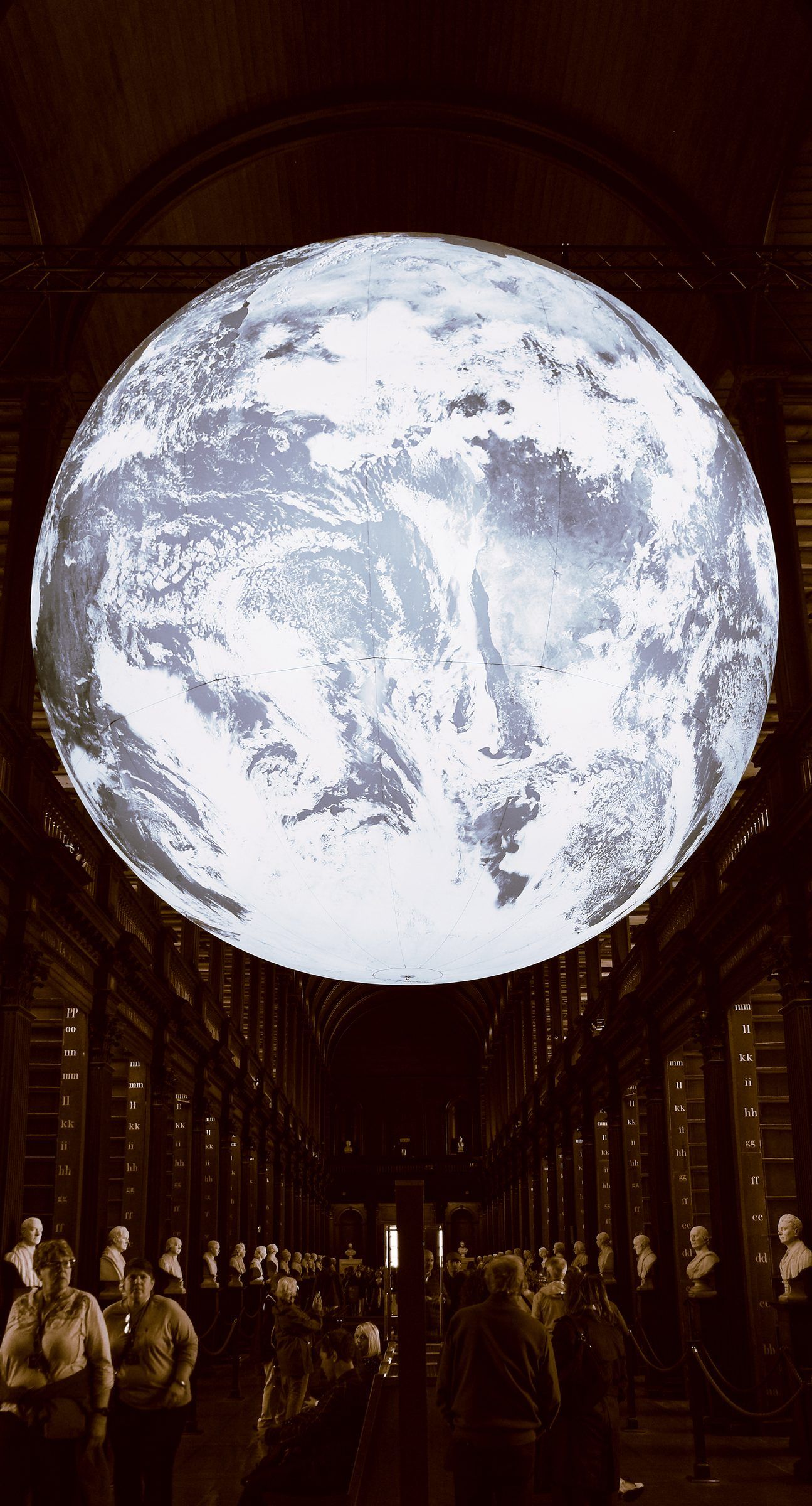 A giant illuminated globe of the Earth hanging in a dark library hall above a crowd of visitors and a row of marble busts