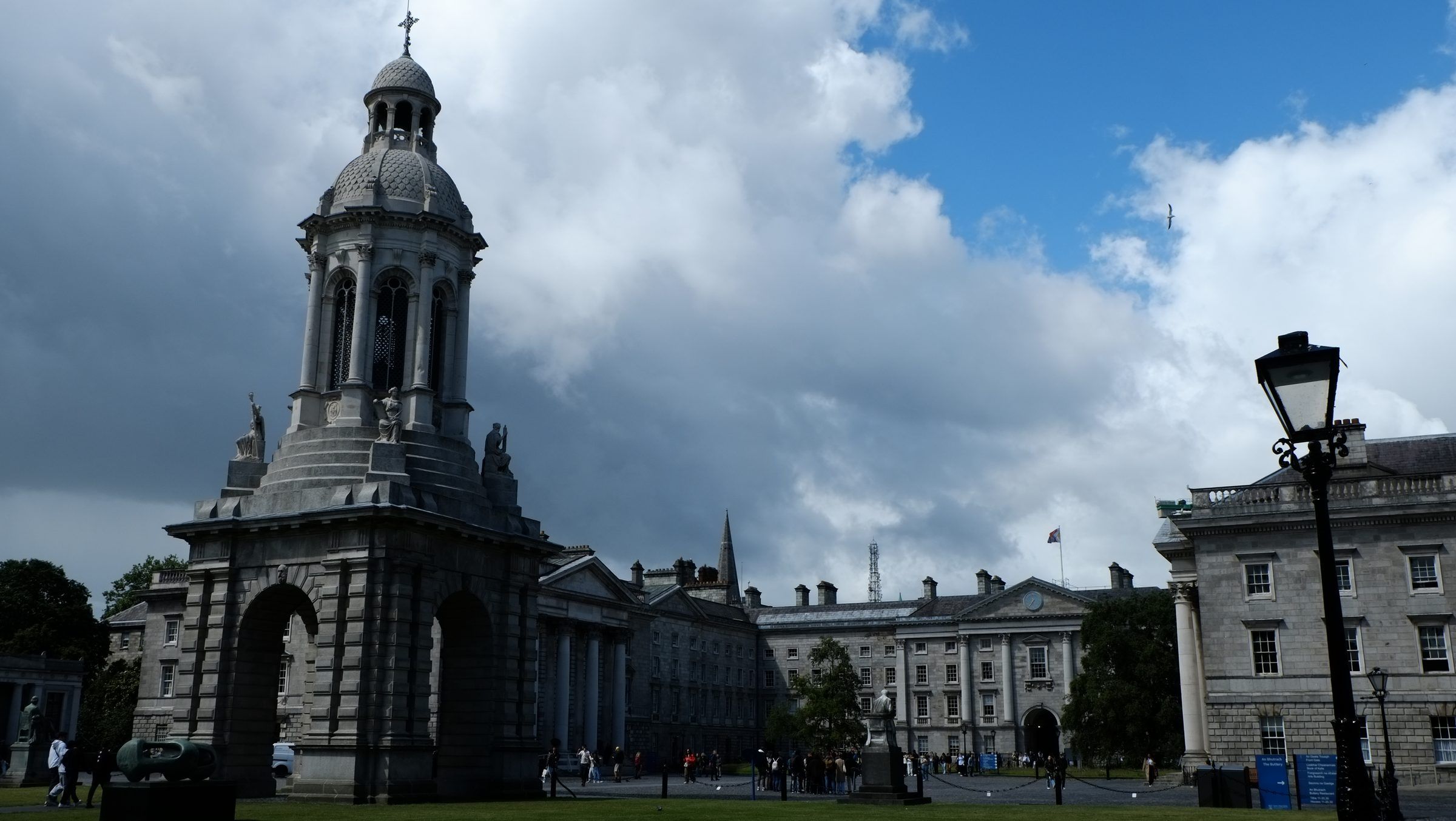 The stone bell tower of Trinity College in shadow beneath dark clouds and a patch of blue sky, an iron lamp post beside it