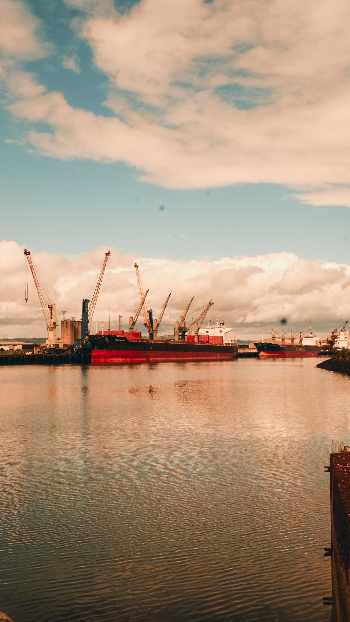 A red cargo ship and dock cranes across still, pink-tinted water under a sky of scattered cloud