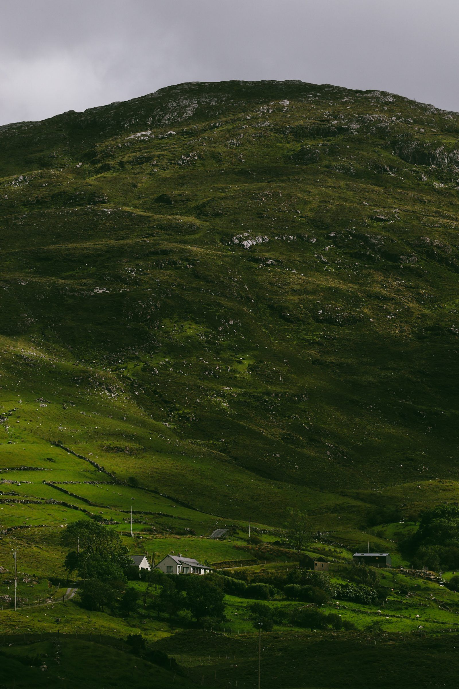 A white cottage among trees at the foot of a vast green mountainside under a grey sky