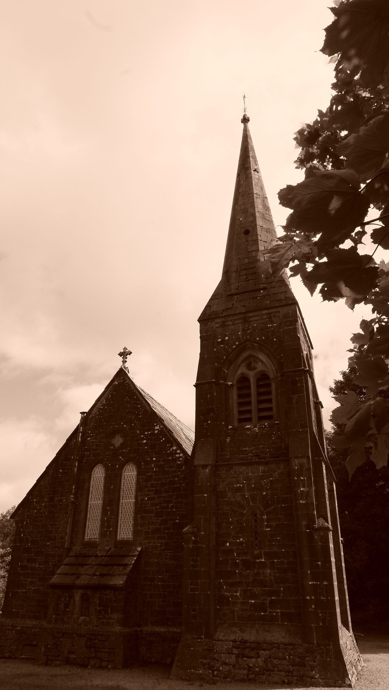 A sepia view of a small stone church with a pointed spire beside a dark tree