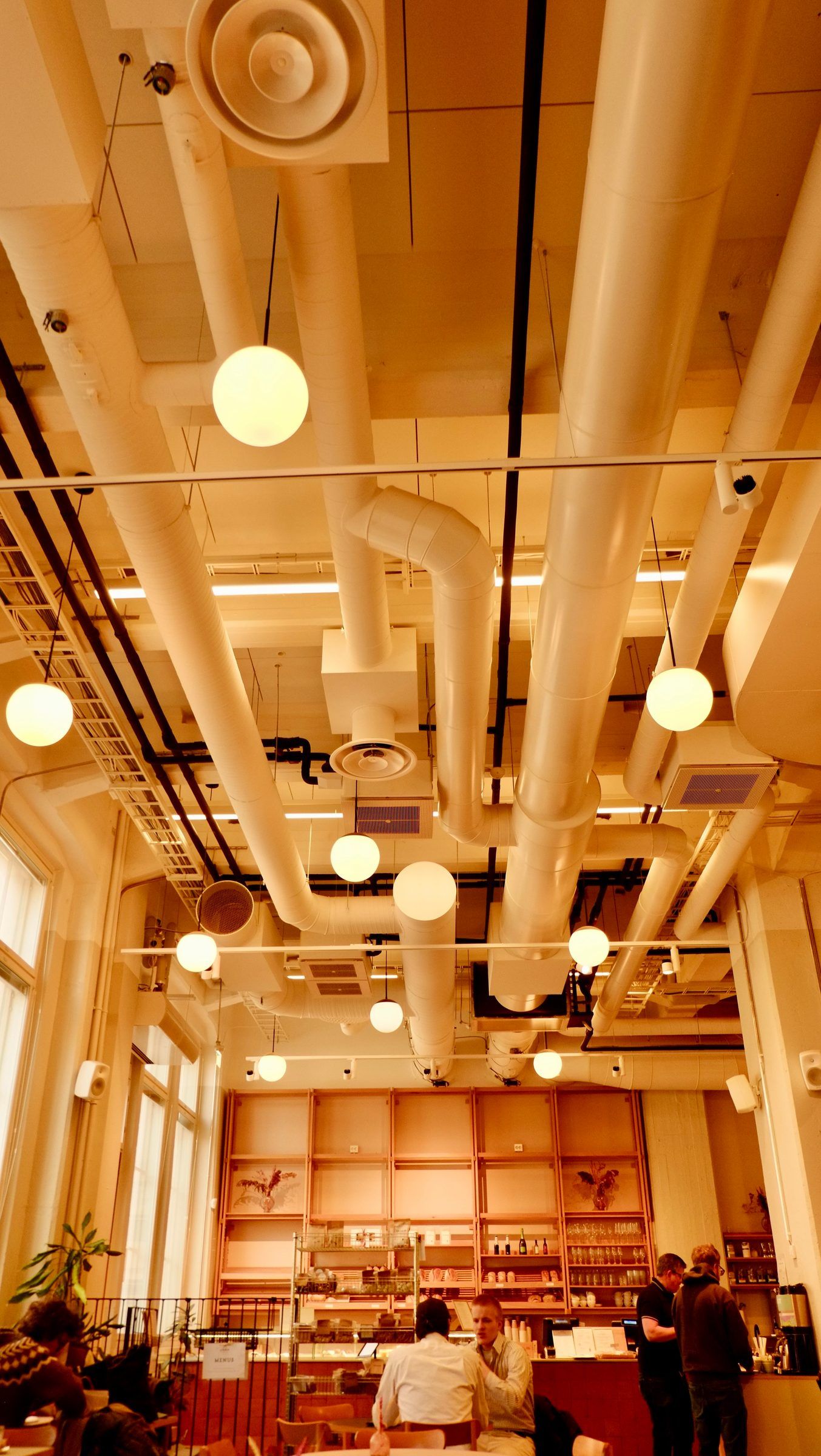 A warm-lit café with white pipes and globe lamps hanging from a high ceiling, people gathered at the counter