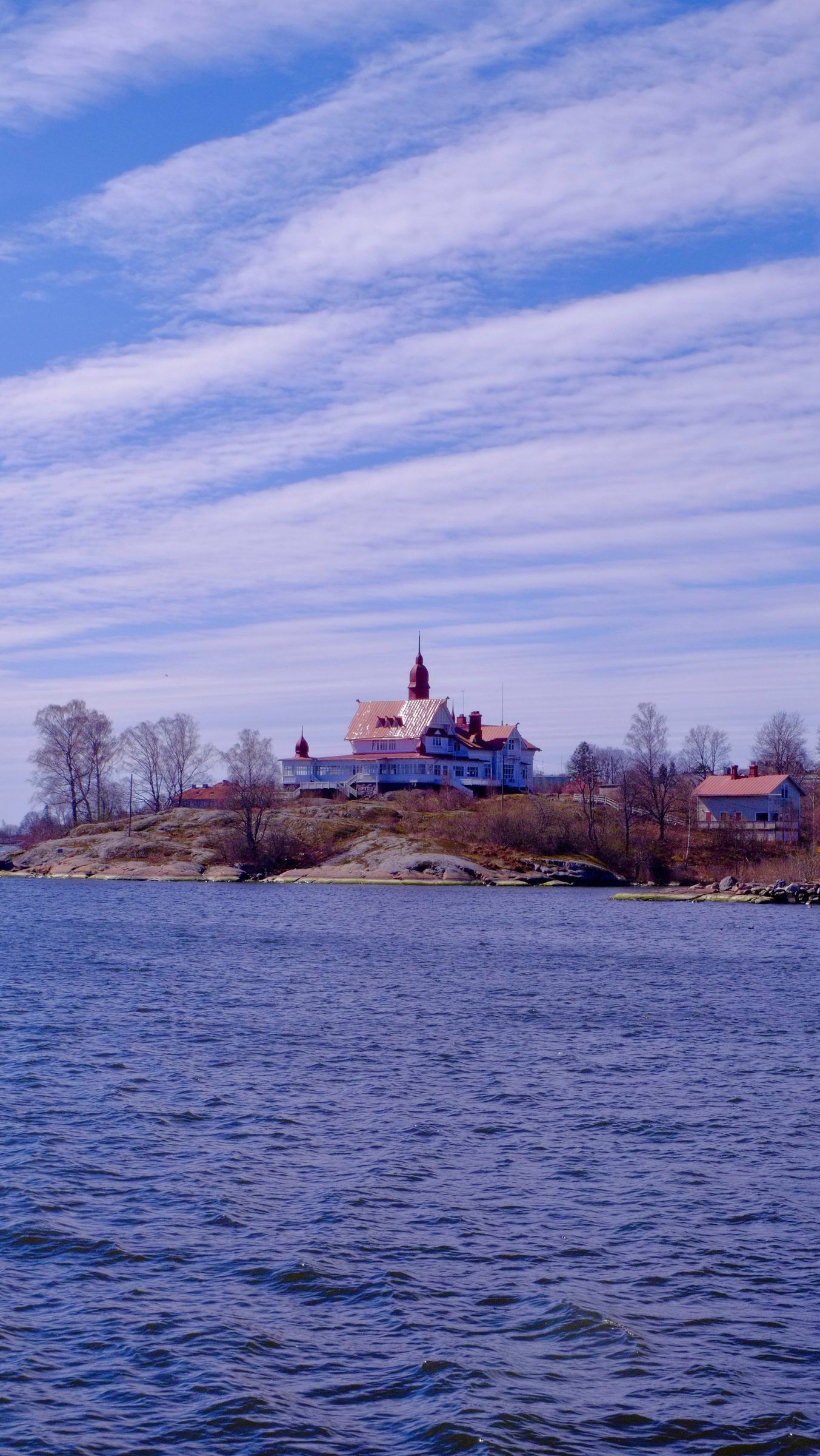 A white villa with a red turret among bare trees on a rocky island, seen across rippling blue water