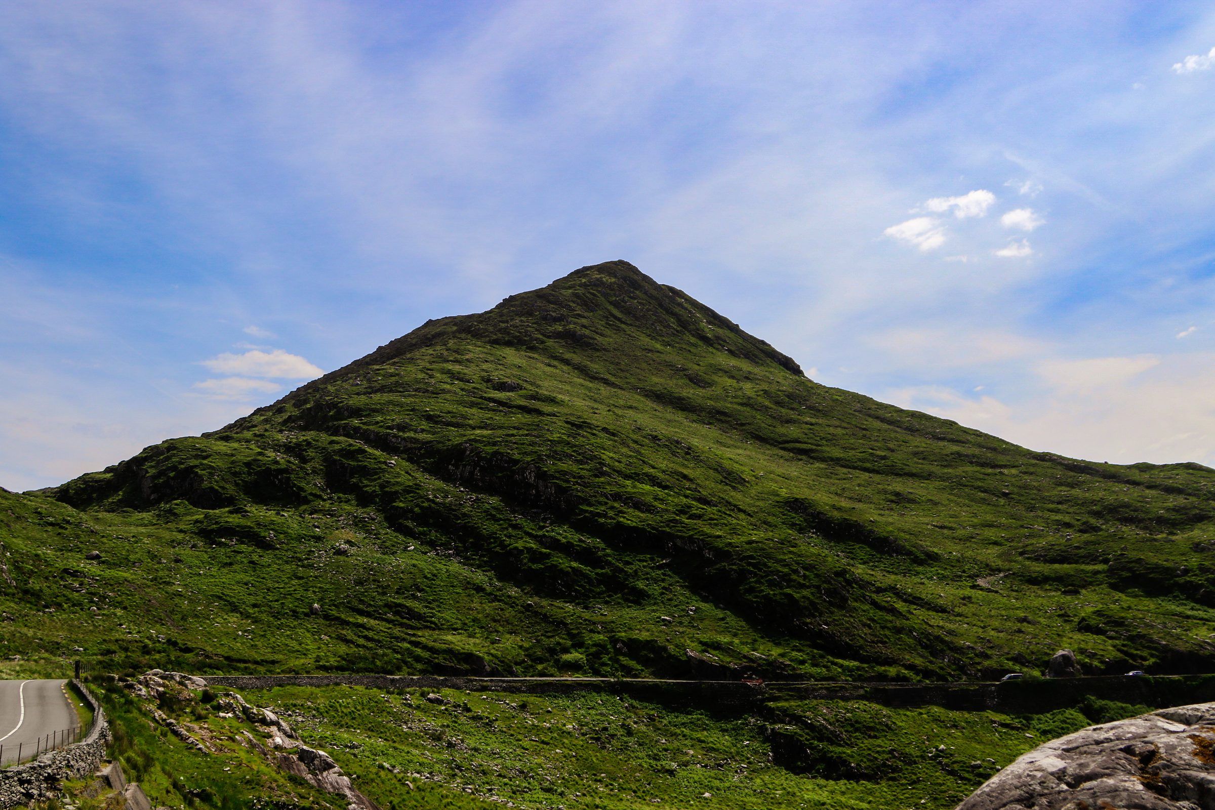 A steep, pointed green hill rising above a mountain road under a streaked blue sky