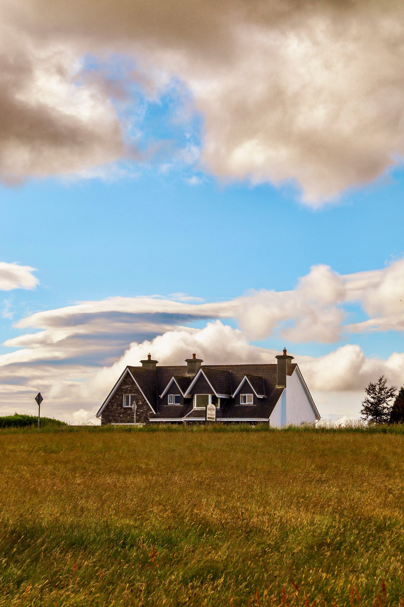 A dark-roofed house with dormer windows standing alone in a golden field beneath billowing clouds