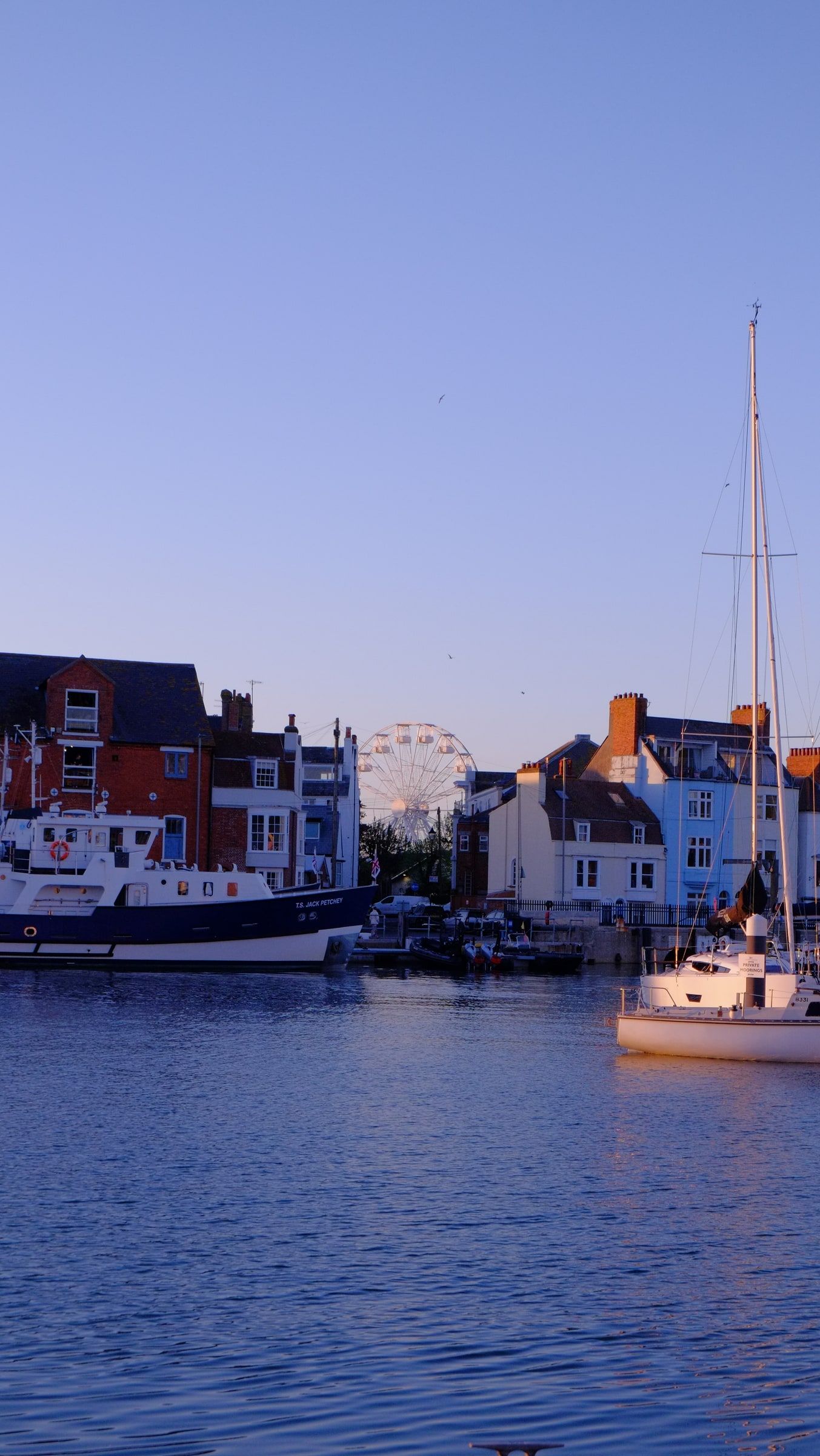 A sailing boat crossing the harbour past painted houses, with a Ferris wheel in the distance