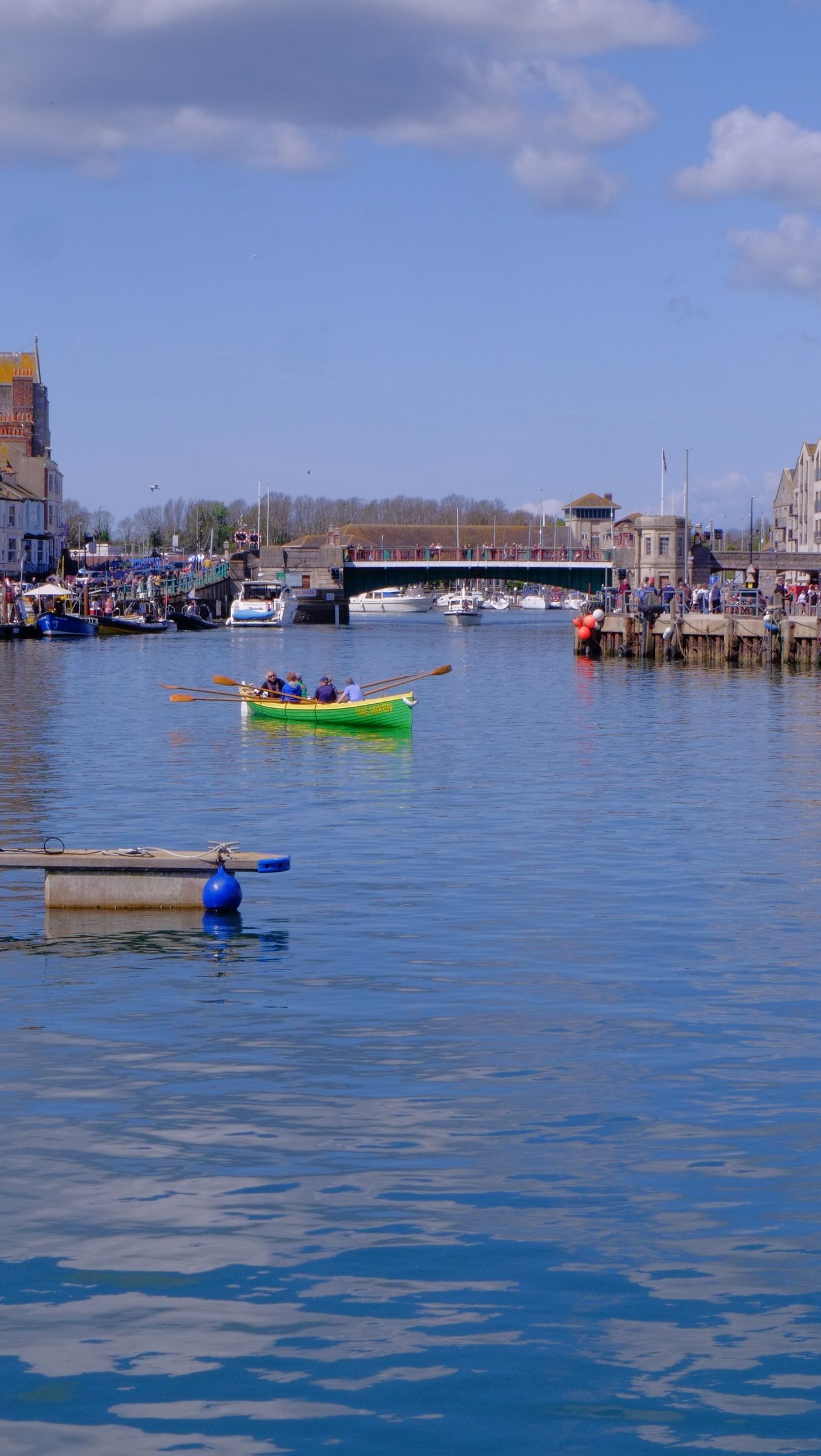 A green rowing boat out on the harbour, the town bridge and crowded quayside in the distance
