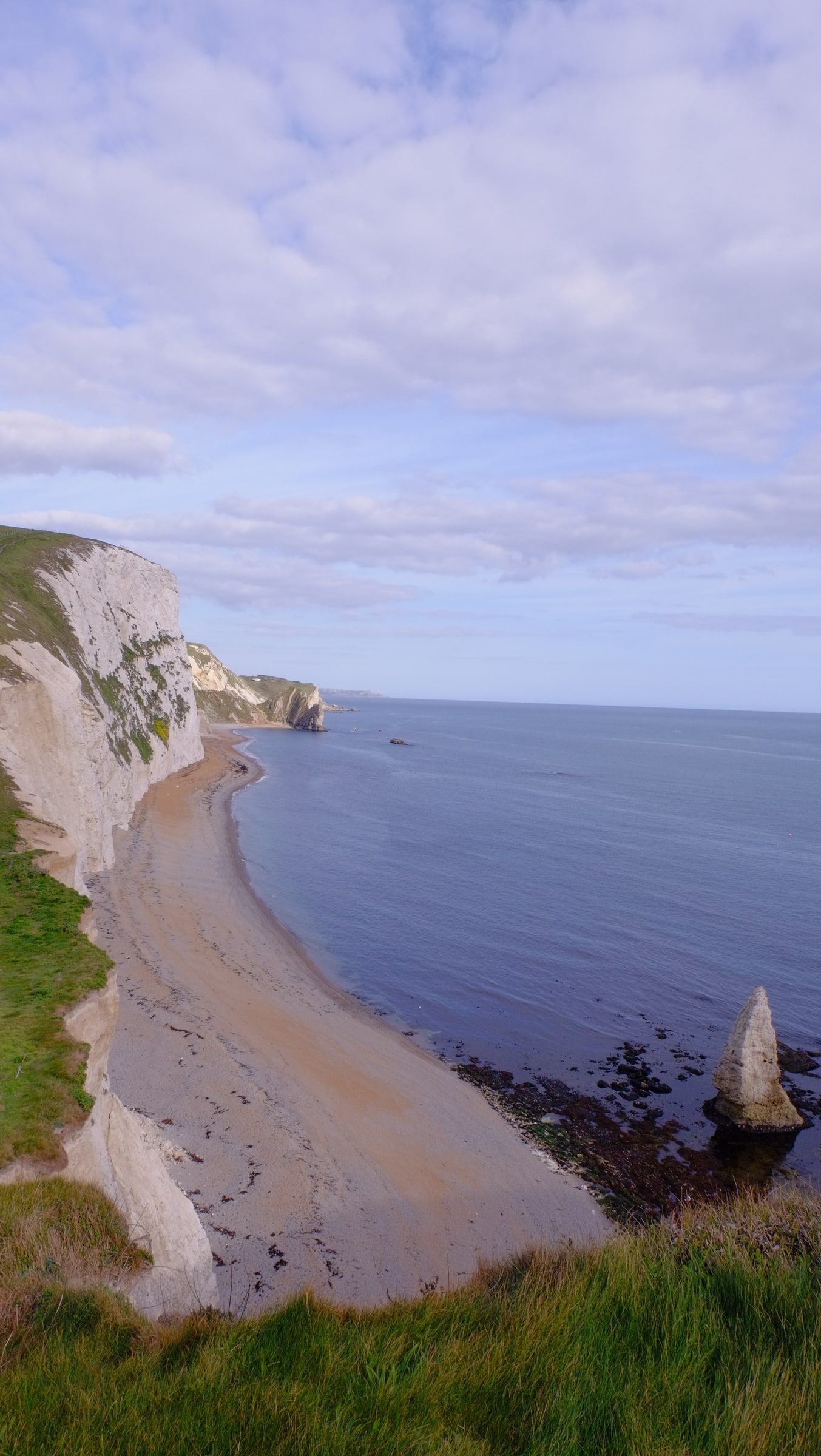 White chalk cliffs curving down to a narrow beach and a sea stack in calm blue water