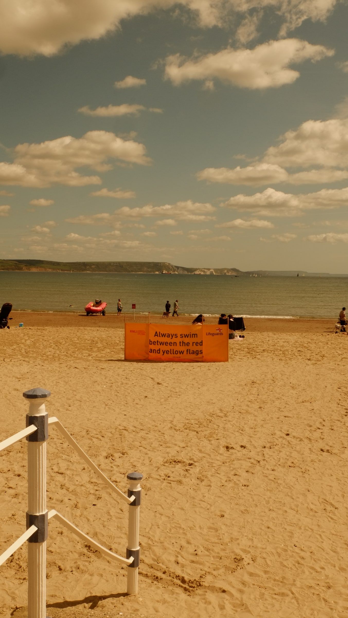 A sandy beach with a roped path, an orange lifeguard sign and people on the shore beneath scattered clouds