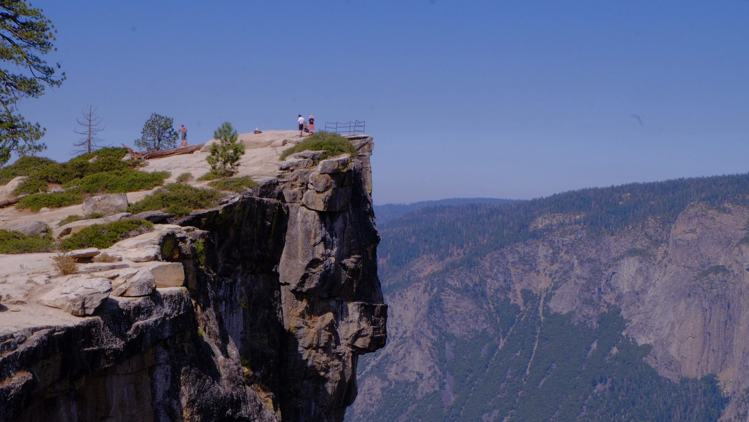 Tiny figures beside a railing on a sheer clifftop overlook, a forested granite canyon wall across the gap